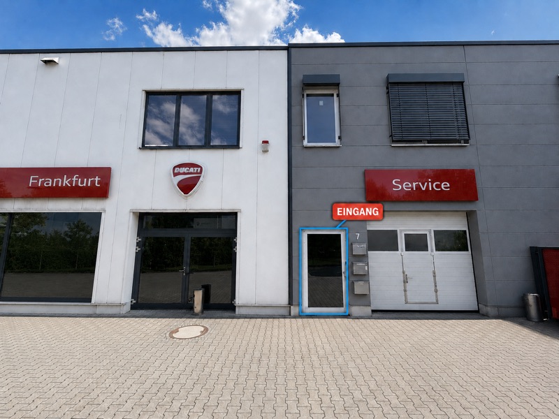 Die Gebäudefront: unsere Eingangstür ist blau umrandet und mit „Eingang“ beschriftet, daneben die Hausnummer 7 mit Briefkasten und Klingel; links davon das Motorradhaus Ducati Frankfurt